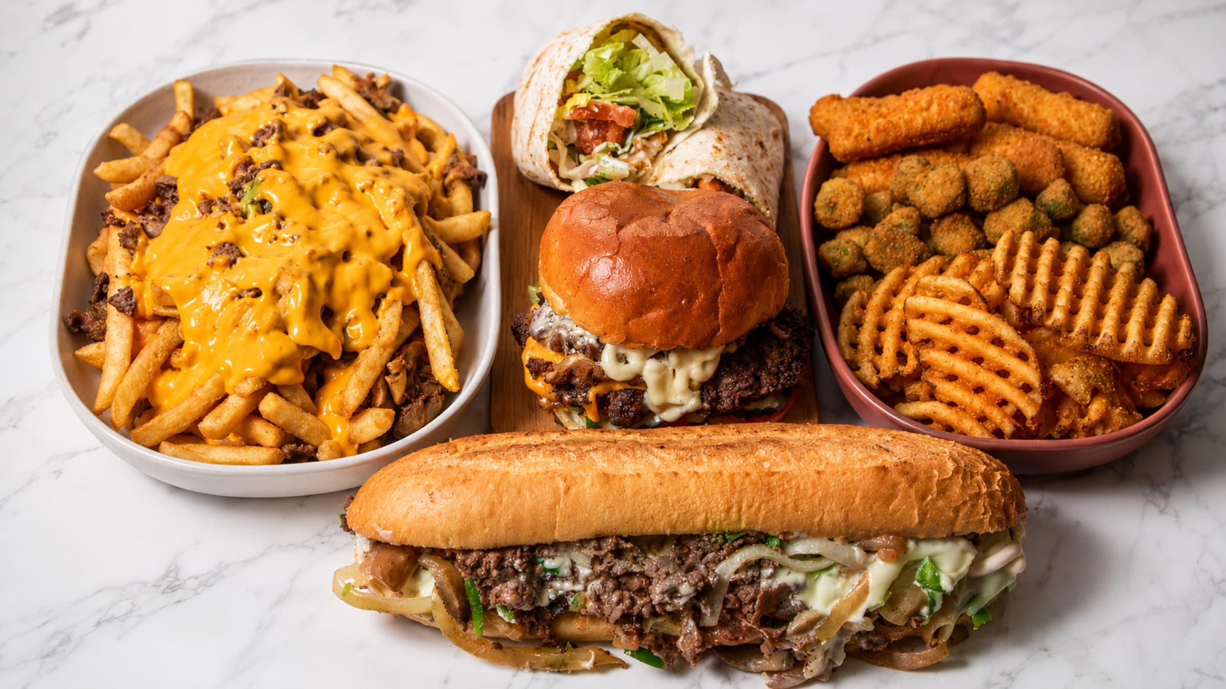 A spread of Moe'z food: loaded cheese fries, a crispy chicken wrap, a stacked double burger, and an Original Philly cheesesteak